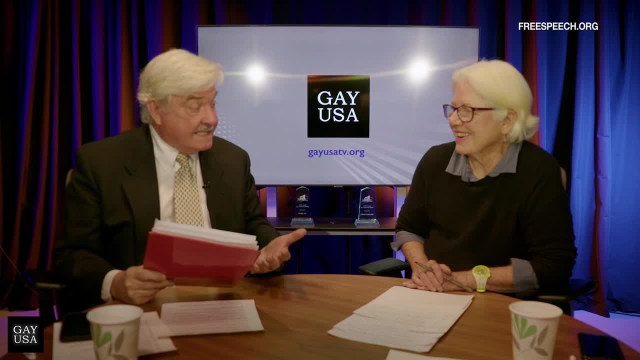 The man in the suit gestures with a red folder towards the woman, who smiles and leans forward.  Behind them, the Gay USA logo is displayed on a screen, with "FreeSpeech.org" in the corner.

