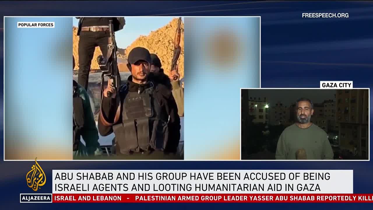 A man in a black cap and vest holds a rifle, looking directly at the camera. On Free Speech TV, a news anchor is reporting from Gaza City.
