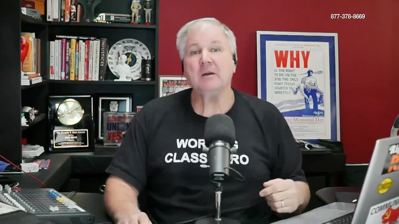 A man with gray hair speaks into a microphone, his words likely broadcast on Free Speech TV. Behind him, a shelf is crammed with books and awards, and a poster with the word "WHY" hangs on the red wall.
