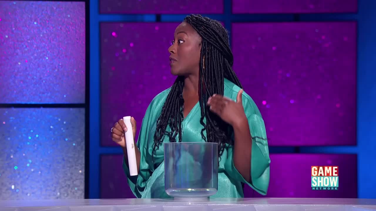 A woman in a teal dress gestures towards a clear glass bowl, holding a white object in her other hand. The Game Show Network logo is visible on the right side of the stage.
