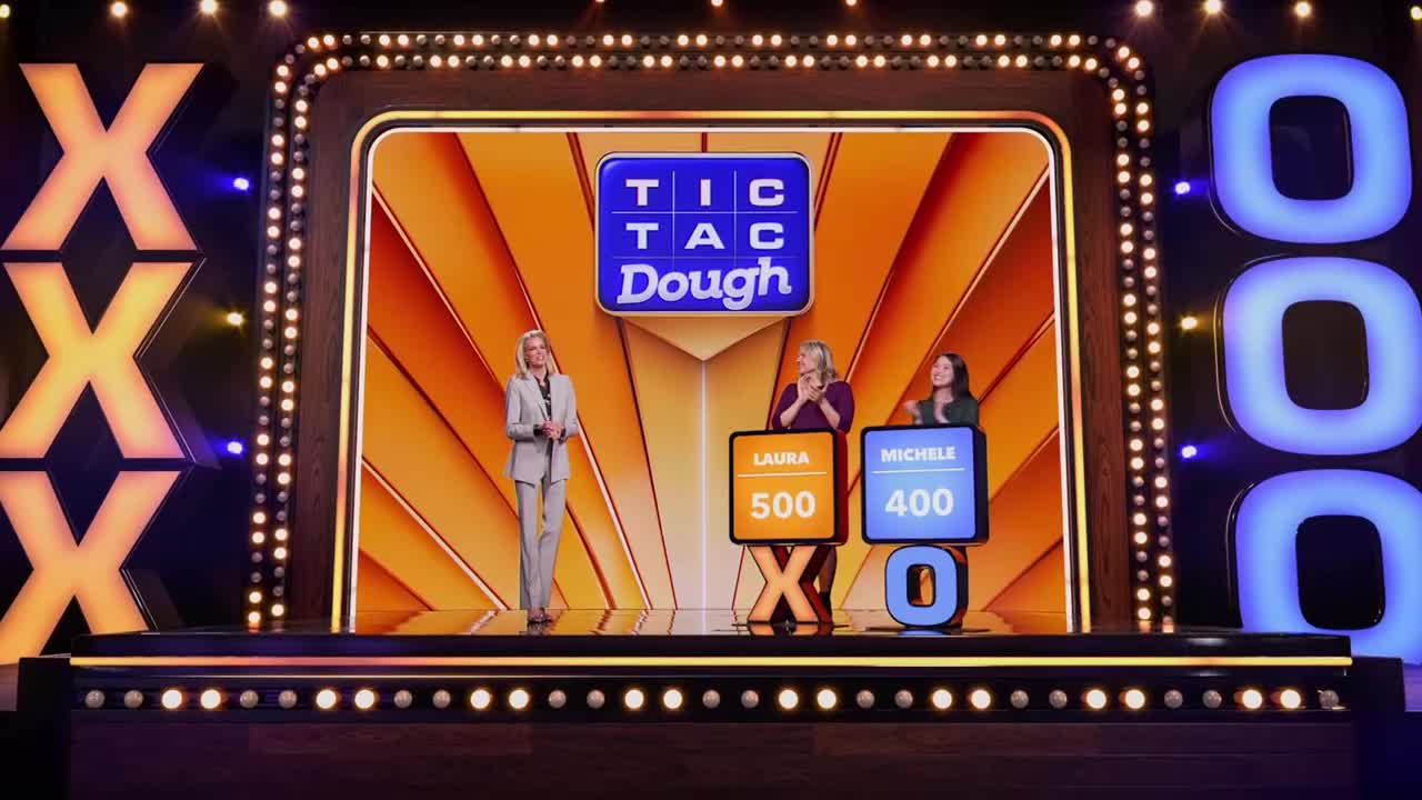 The host stands center stage, while two contestants flank her with their scoreboards. The "Tic Tac Dough" set is lit up, ready for the next round.
