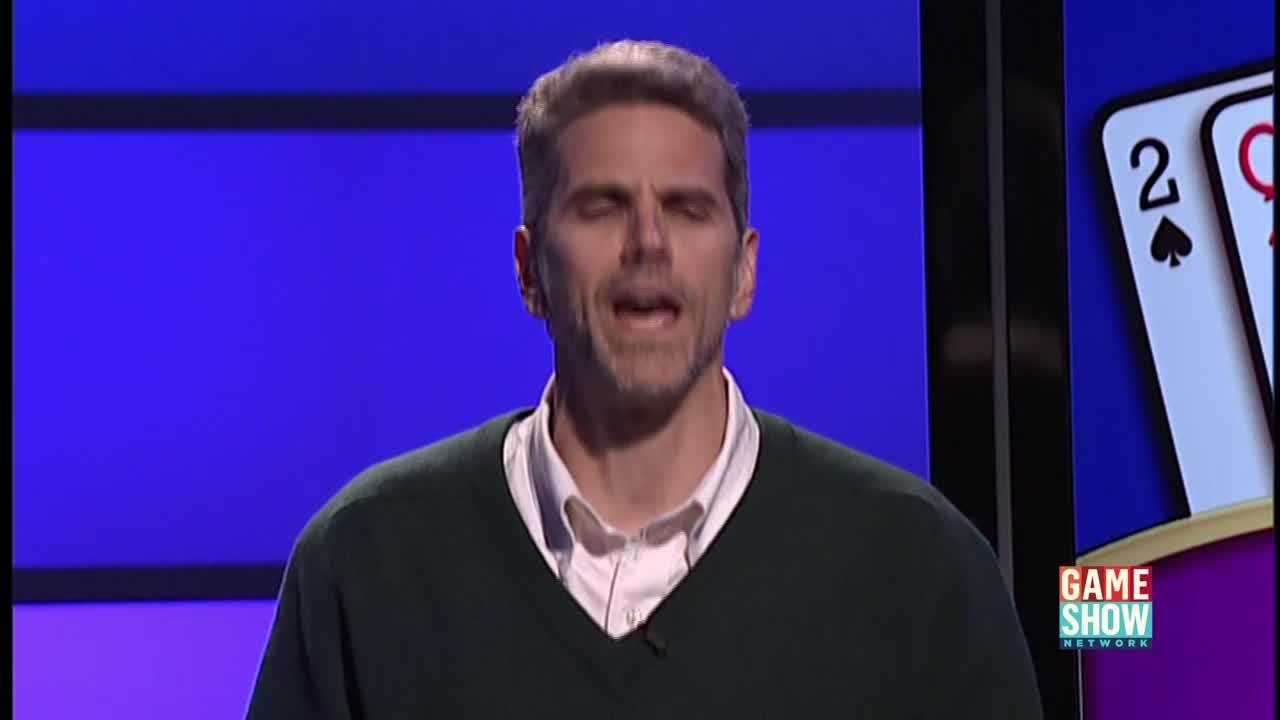 The man in the green sweater is mid-sentence, his mouth open as he speaks. Behind him, the Game Show Network logo and playing cards are visible on a screen.
