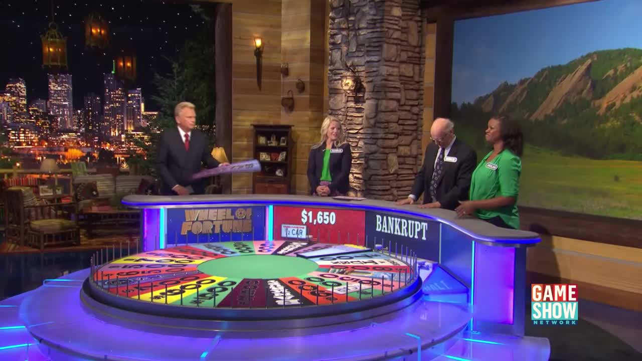 The host, holding a clue, gestures toward the "Wheel of Fortune" wheel. The contestant in green stands, looking on, as the wheel spins.
