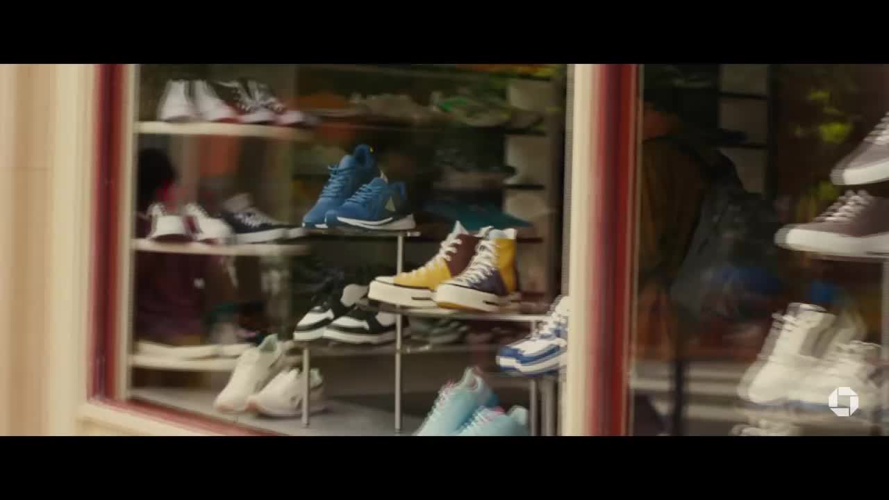 A shoe store window displays rows of sneakers, from high-tops to low-cuts, in a variety of colors. The storefront is slightly blurred, suggesting movement, perhaps a passing vehicle.
