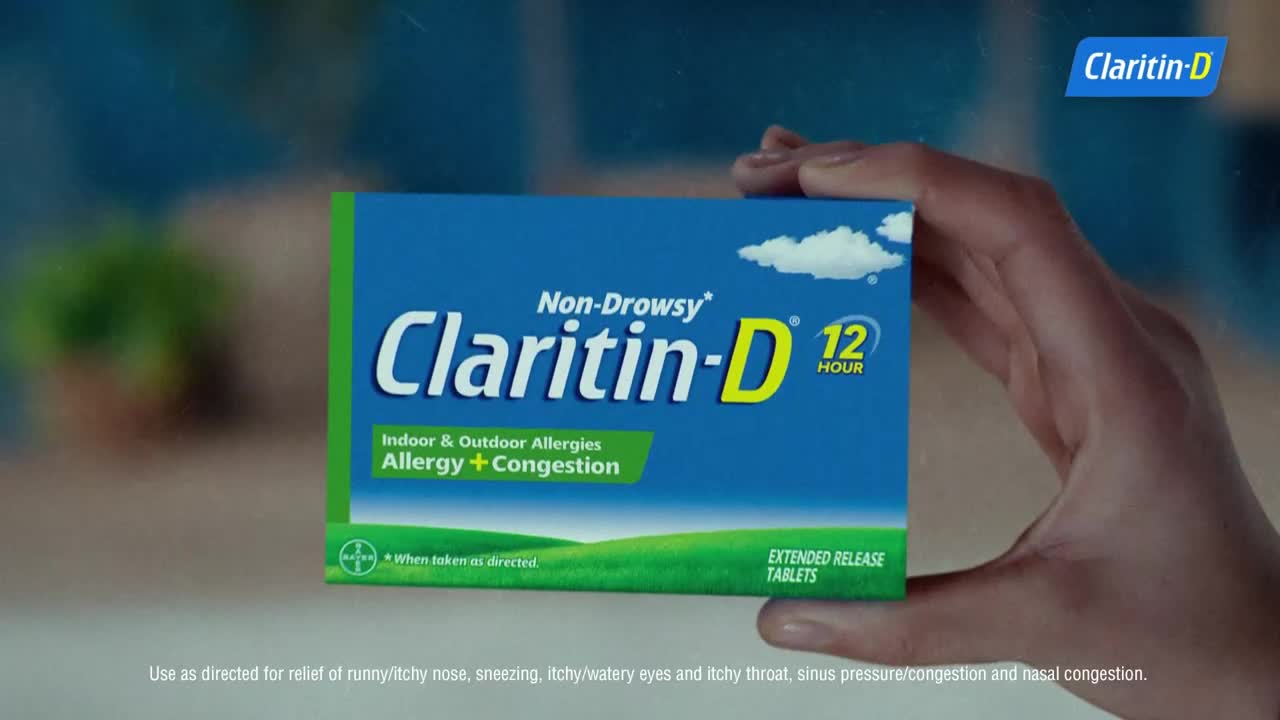 A hand presents a box of Claritin-D, its blue background contrasting with the green and white details. The box is held up against a blurred background, likely a home setting.
