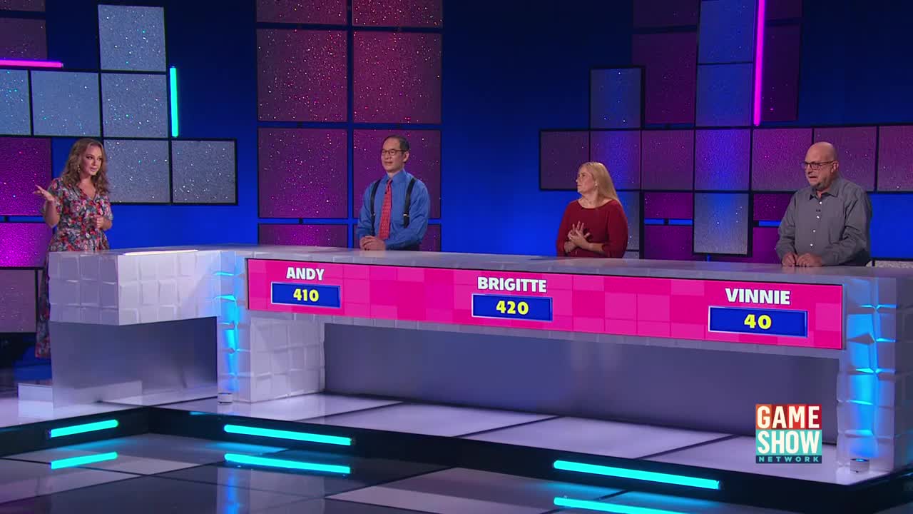 A woman in a floral dress gestures to the three contestants seated behind a long, pink desk. Each person's name and score are displayed on a screen in front of them, as the Game Show Network logo sits in the corner.
