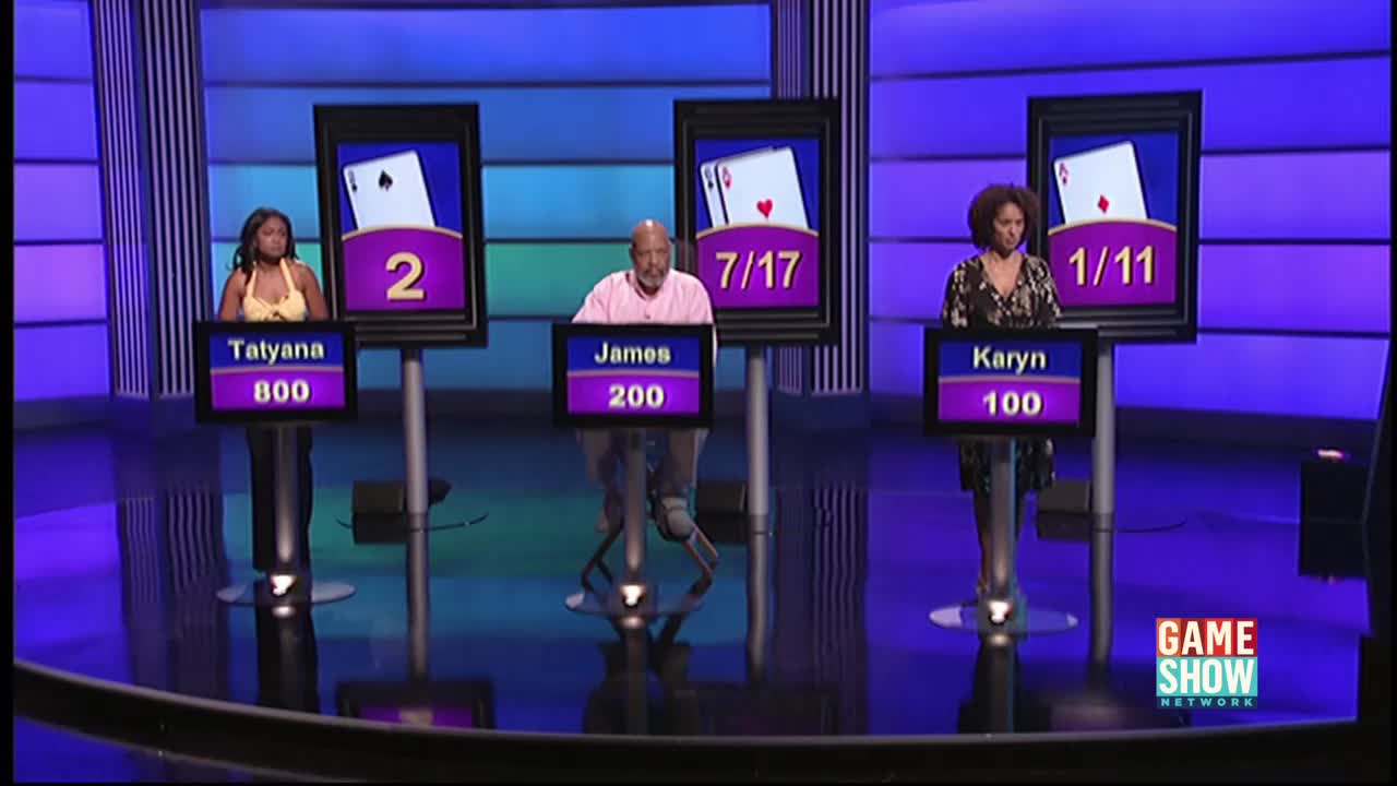 Three contestants stand behind podiums on a Game Show Network set, each facing a screen displaying playing cards and a score. James sits on a stool between Tatyana and Karyn, all poised to answer questions.
