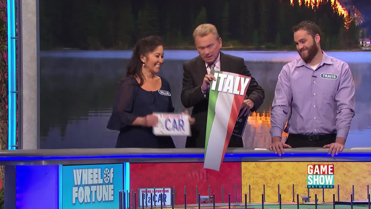 The host is holding up a giant "Italy" card, while the woman to his left clutches a "Car" prize card. A contestant named Travis stands to the right, smiling.
