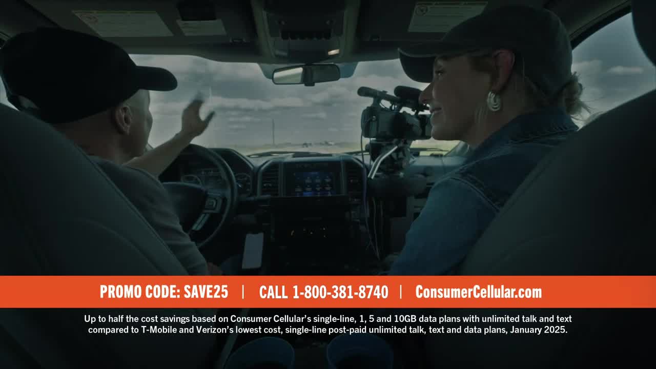 A man in a baseball cap gestures with his right hand while driving. Across from him, a woman wearing a denim jacket and a cap smiles, a camera mounted on a tripod between them.
