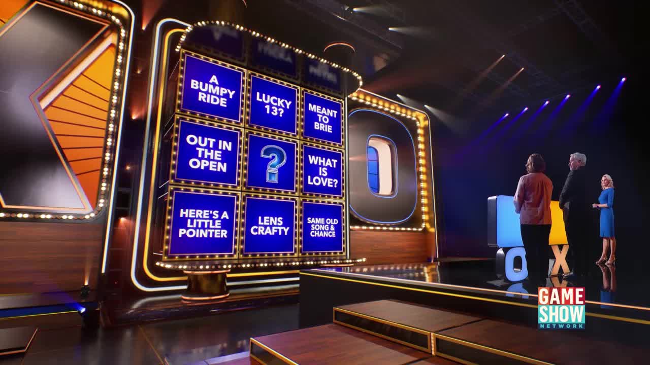 A large, illuminated game board with multiple categories is lit up on a stage. Three contestants stand on a platform, facing the board, while a host in a blue dress looks on.
