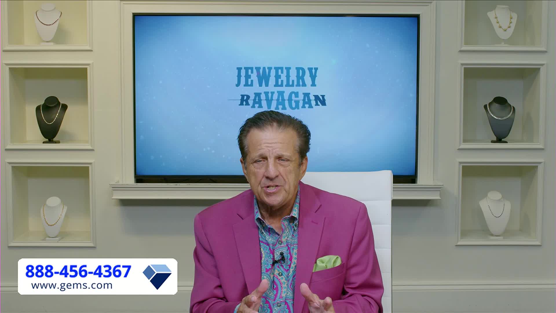 A man in a bright pink jacket gestures with his hands while speaking, the "Jewelry Ravagan" logo behind him.  Necklaces are displayed on mannequins in the wall, and a Gem Shopping Network phone number is visible at the bottom.
