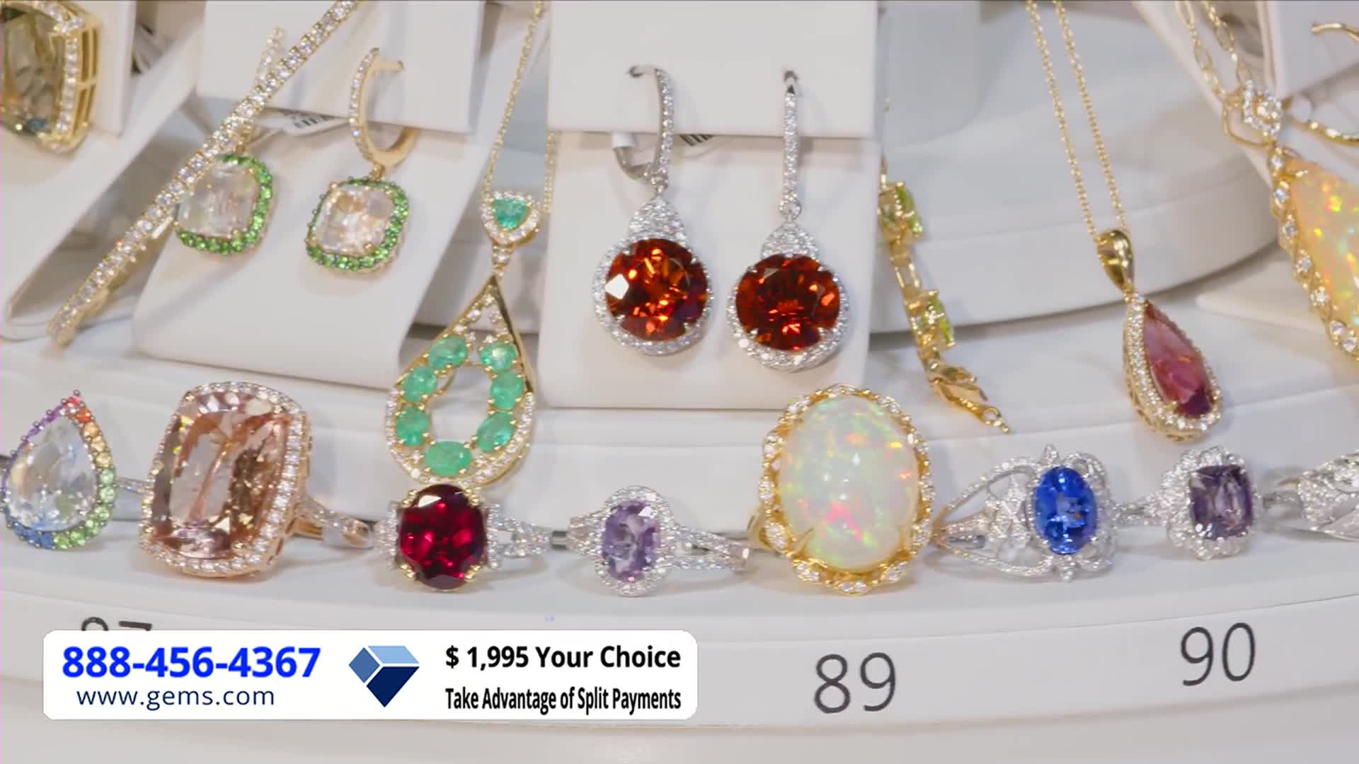 A rotating display showcases a collection of jewelry, with rings and pendants sparkling under bright lights. The Gem Shopping Network logo and a phone number are visible in the corner, advertising a "Your Choice" deal.
