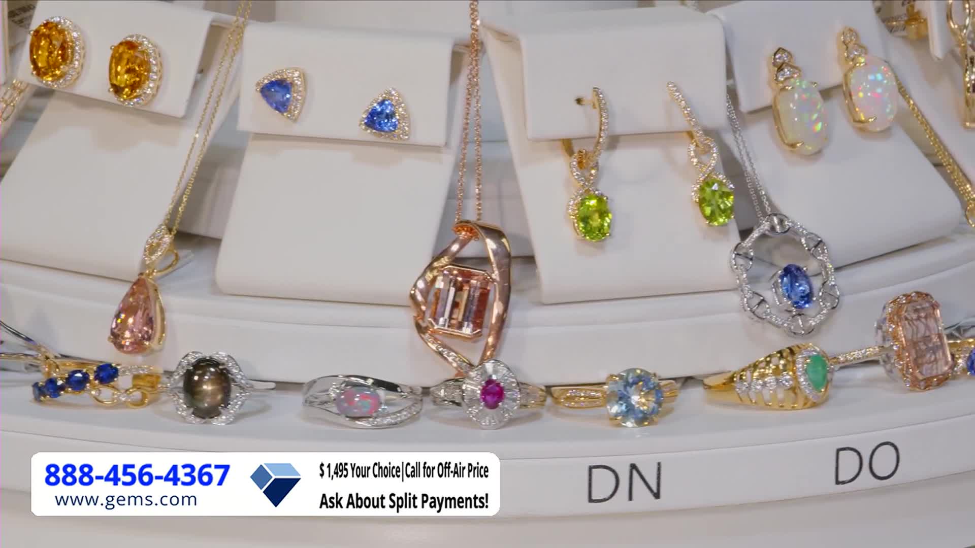 Sparkling jewelry is arranged on white display stands, with rings and necklaces catching the light. A Gem Shopping Network call-in number and website are visible in the lower left corner.
