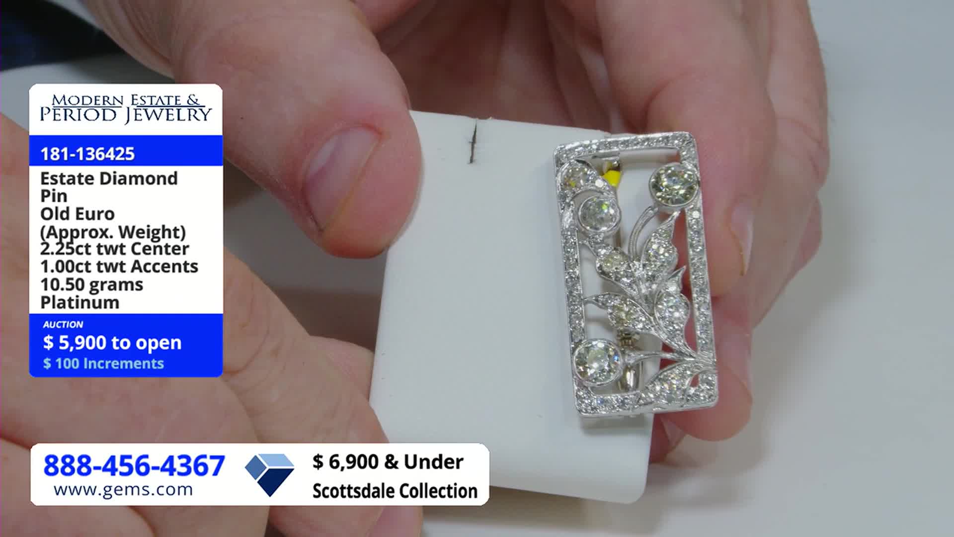 A hand holds a platinum and diamond pin, showcasing it against a white backdrop. The Gem Shopping Network logo and auction details are visible in the corner.
