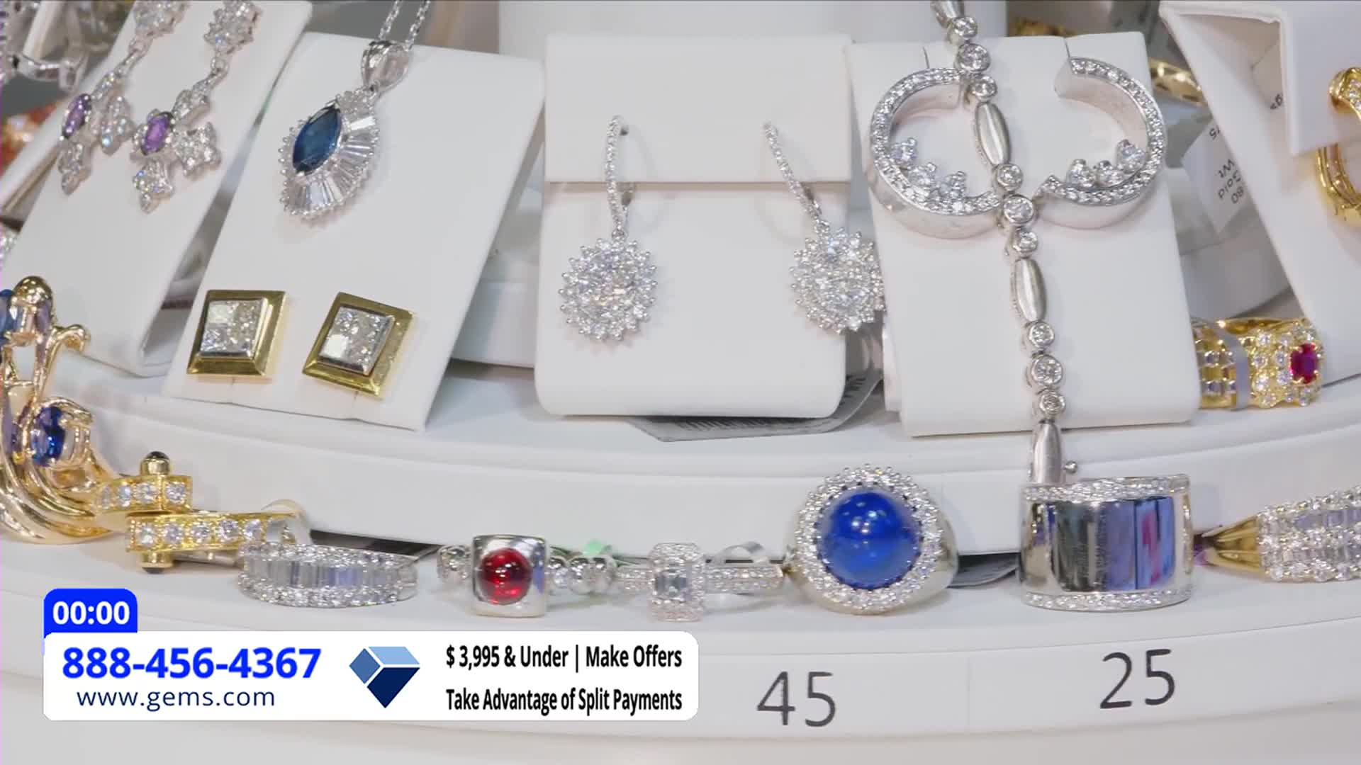 Sparkling jewelry, displayed on white pedestals, fills the frame. A ring with a large blue stone sits prominently, while the Gem Shopping Network logo and phone number are visible in the corner.
