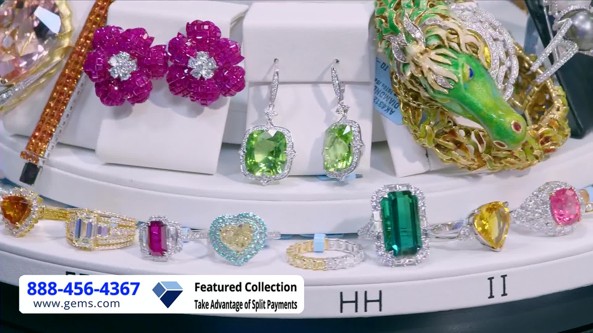 On a white surface, several pieces of jewelry are arranged, including earrings with green stones and rings with various colored gems. A call-to-action graphic for Gem Shopping Network appears at the bottom of the screen.
