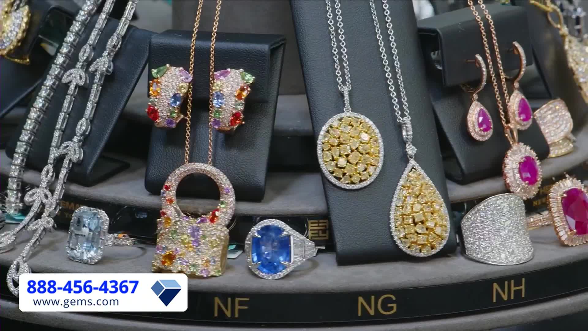 Sparkling jewelry, including necklaces and rings, is arranged on a rotating display. The Gem Shopping Network logo and phone number are visible in the bottom left corner.
