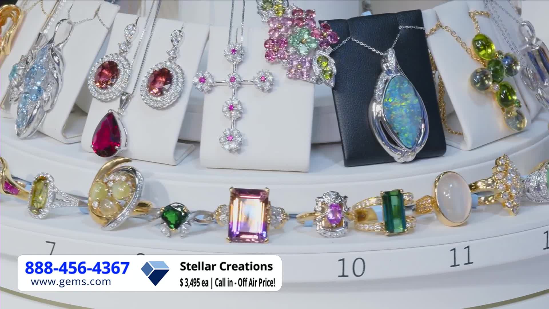 Jewelry, displayed on white stands and a rotating platform, sparkles under bright lights. A phone number and website for Gem Shopping Network are displayed in the corner.
