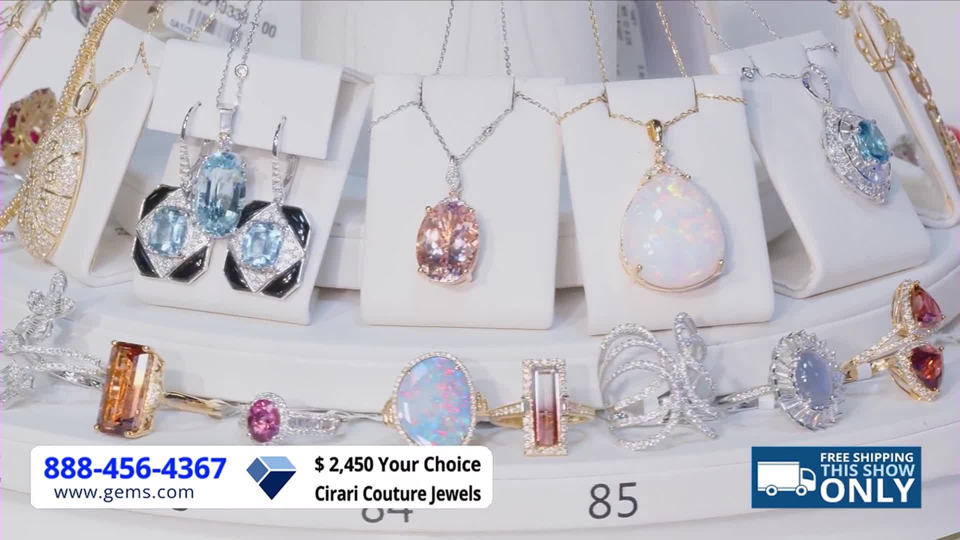 Jewelry, including necklaces and rings, sits on display, illuminated by bright lights. The Gem Shopping Network logo and a phone number are visible at the bottom of the screen.

