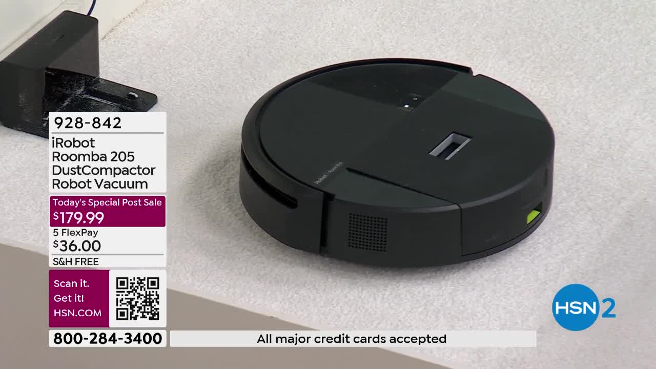 A black iRobot Roomba 205 sits on a light-colored carpet. The vacuum has a low profile and a circular shape, with a charging dock to its left. The HSN2 logo is visible in the lower right corner.