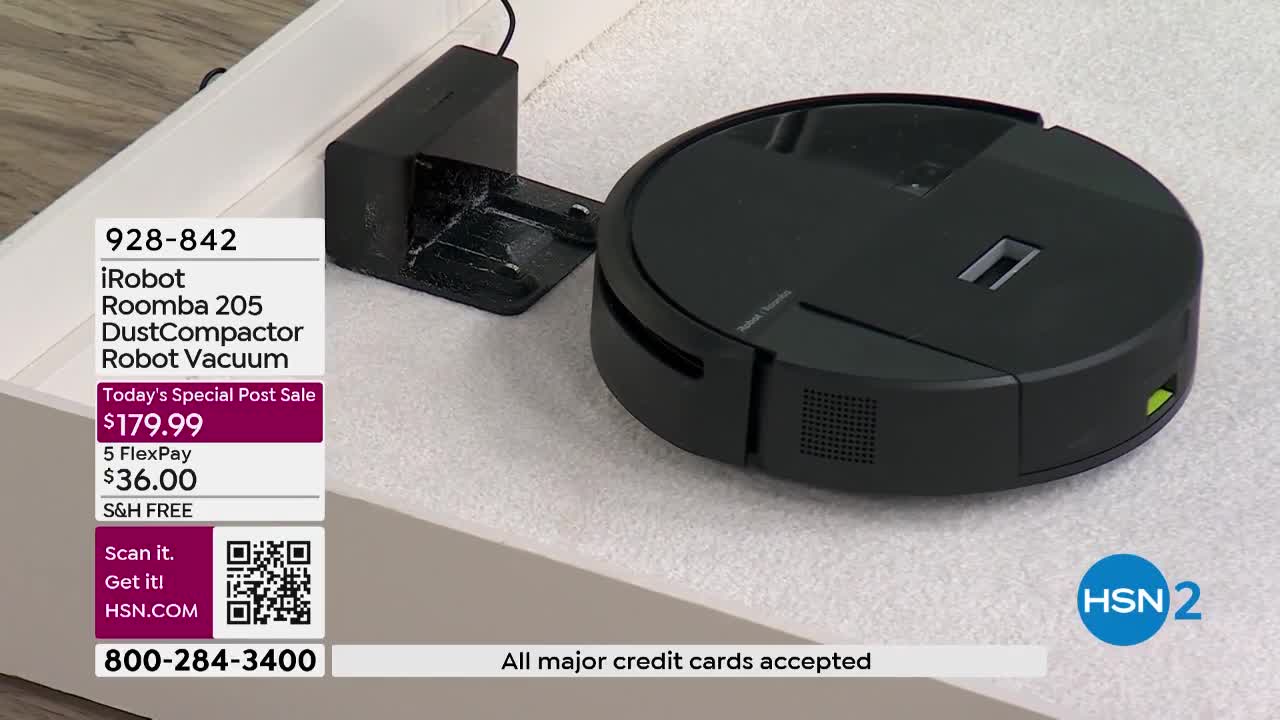 The black iRobot Roomba 205 sits on a textured surface next to its charging dock. A green light glows on the side of the vacuum.