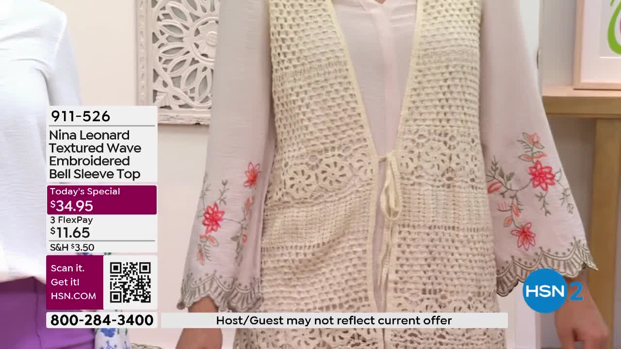 A woman stands on a set, wearing a cream-colored crocheted vest over a white bell-sleeved top. The top is adorned with delicate floral embroidery in shades of pink and green. A graphic overlay displays product information and a phone number for HSN.