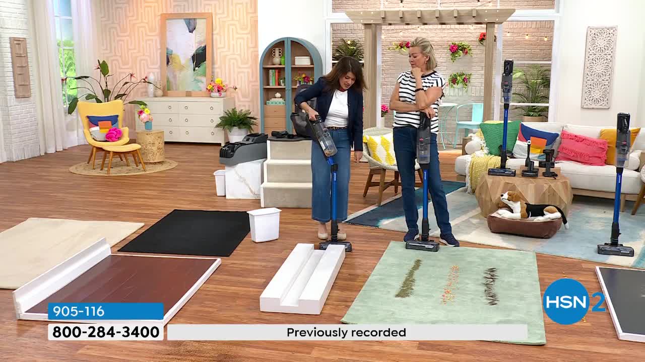 A woman in a dark jacket and jeans demonstrates a blue stick vacuum on a white ramp. Another woman, in a striped shirt and jeans, stands nearby with her own blue vacuum.