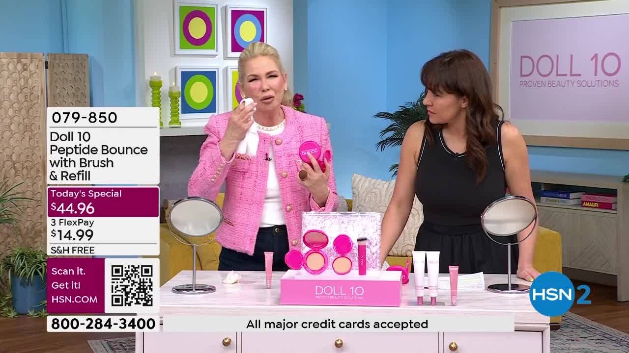 A woman in a pink jacket dabs her face with a white puff while holding a compact. Beside her, another woman in a black dress watches the demonstration.