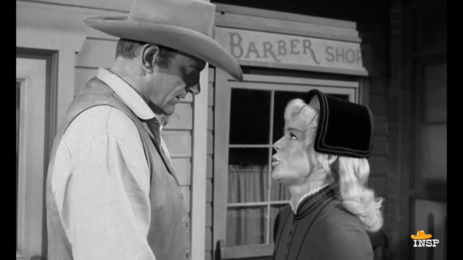 A man in a cowboy hat and vest stares down at a woman in a bonnet, both standing outside a barber shop. She's looking up at him, her mouth slightly open as if she's speaking.
