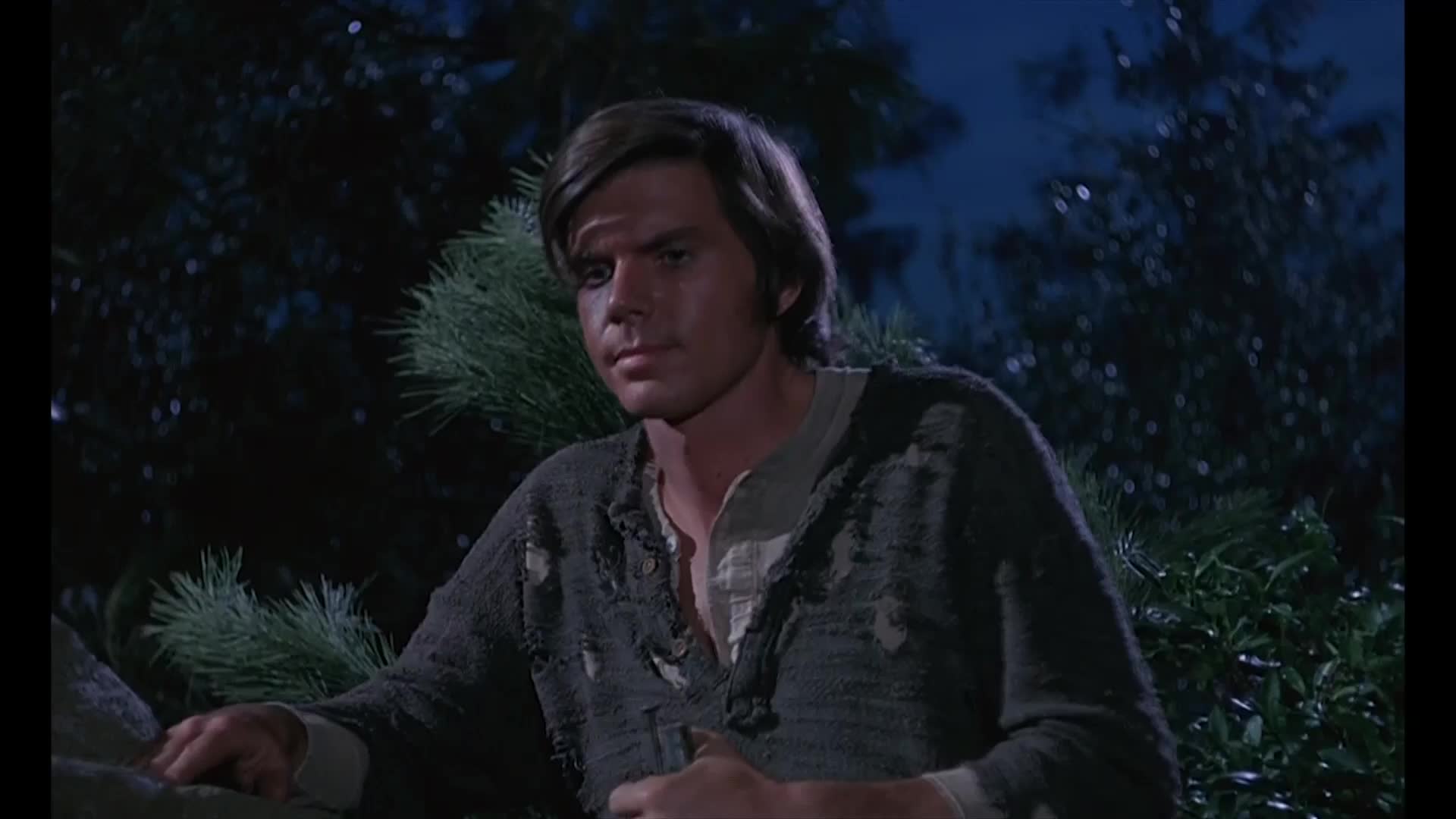 The man is looking down, his gaze thoughtful, framed by dark trees. He's wearing a tattered gray shirt over a white one, the scene lit by the soft glow of a night sky.
