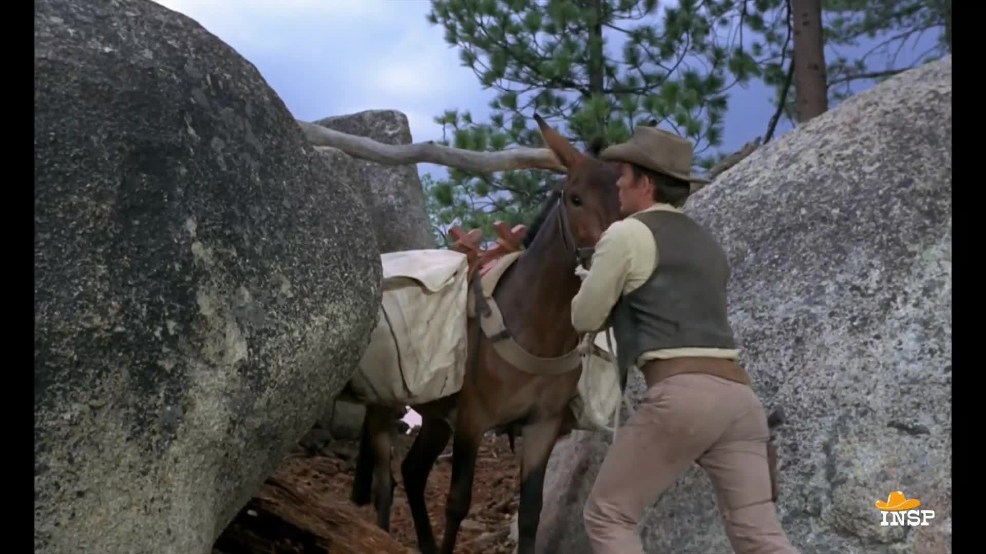 A man in a cowboy hat is nudging a pack mule between two large boulders. The animal is loaded down with saddlebags, and the man is pushing it forward.
