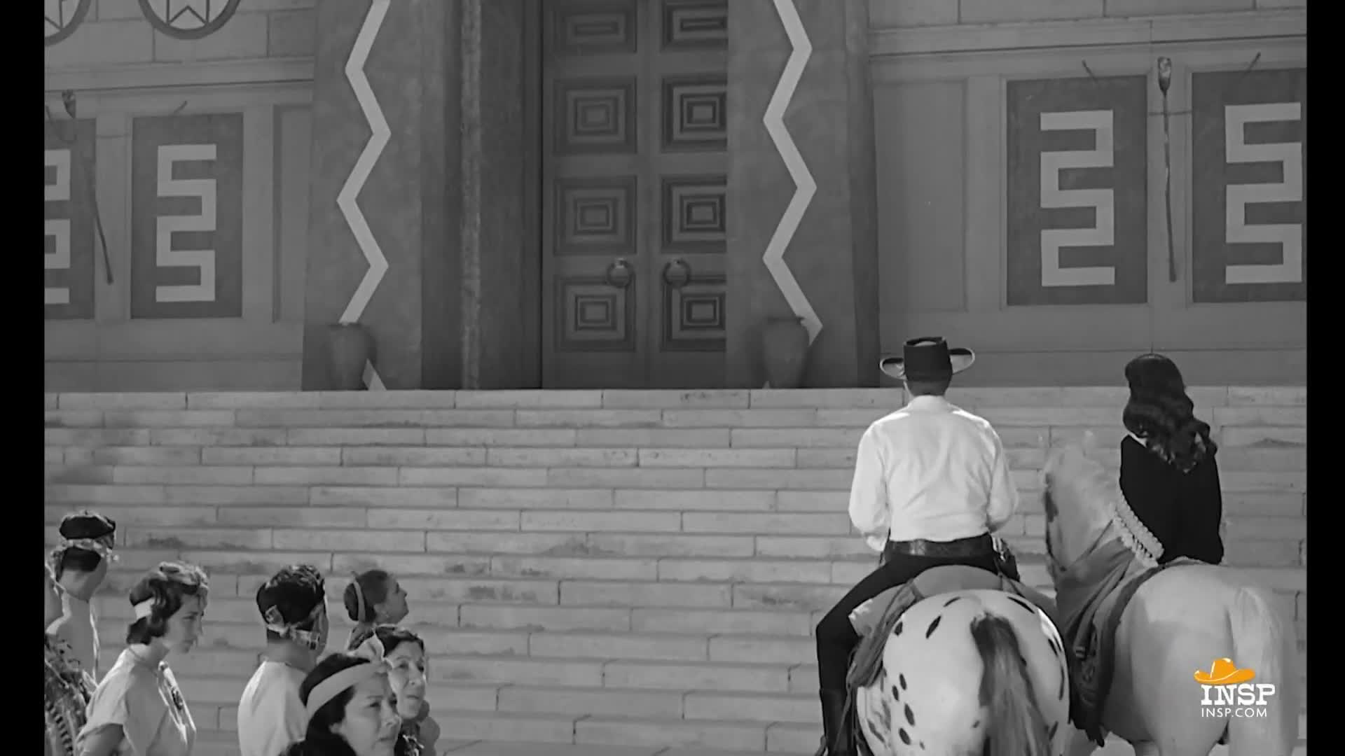 A man in a cowboy hat sits atop a spotted horse, facing a large wooden door. Several people stand below, watching as he and another rider on a white horse prepare to move.

