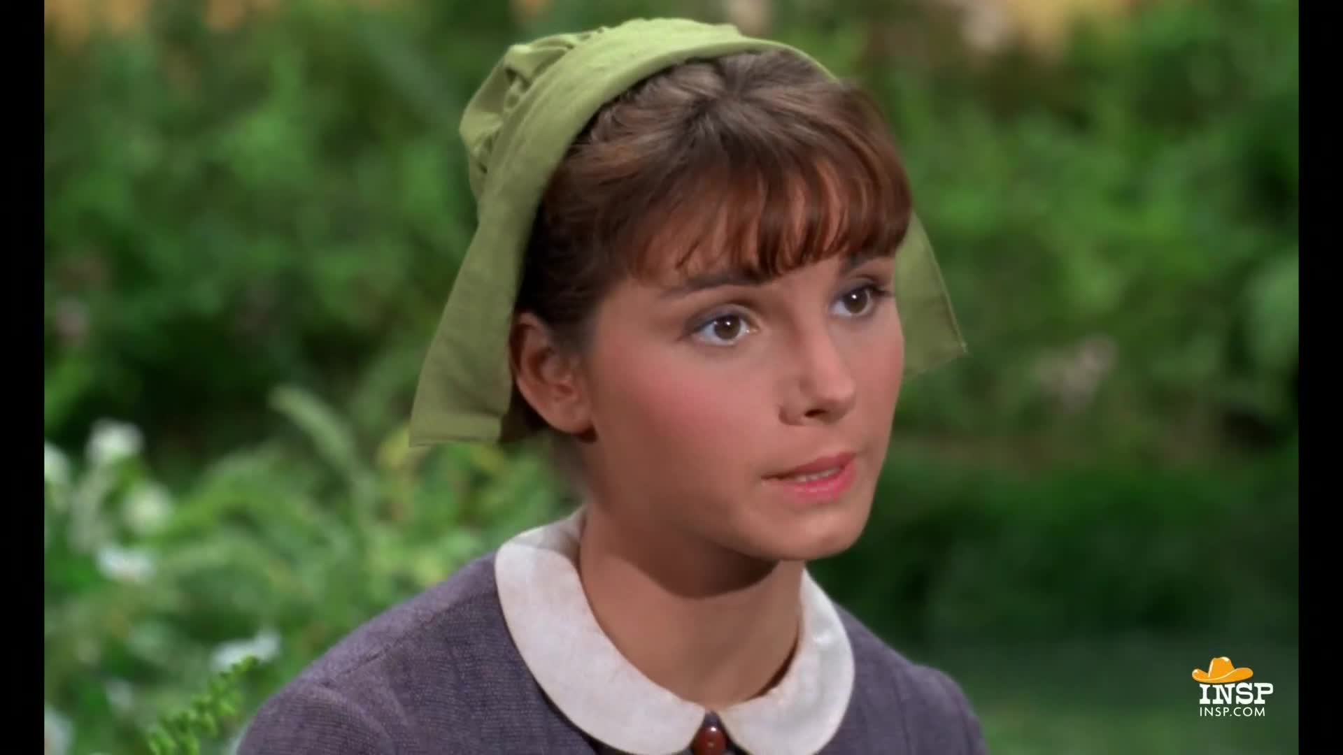 Her eyes are fixed on something off-camera, and her lips are slightly parted as if she's about to speak. A green headscarf frames her face, and a white-collared dress suggests a scene from a show on INSP.
