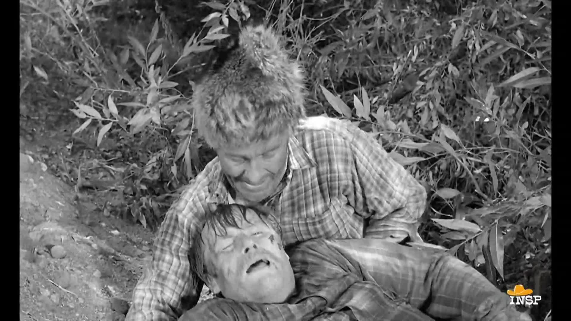 A man in a fur hat leans over another man lying on the ground, his face showing concern. The man on the ground looks up, his eyes wide and mouth open.
