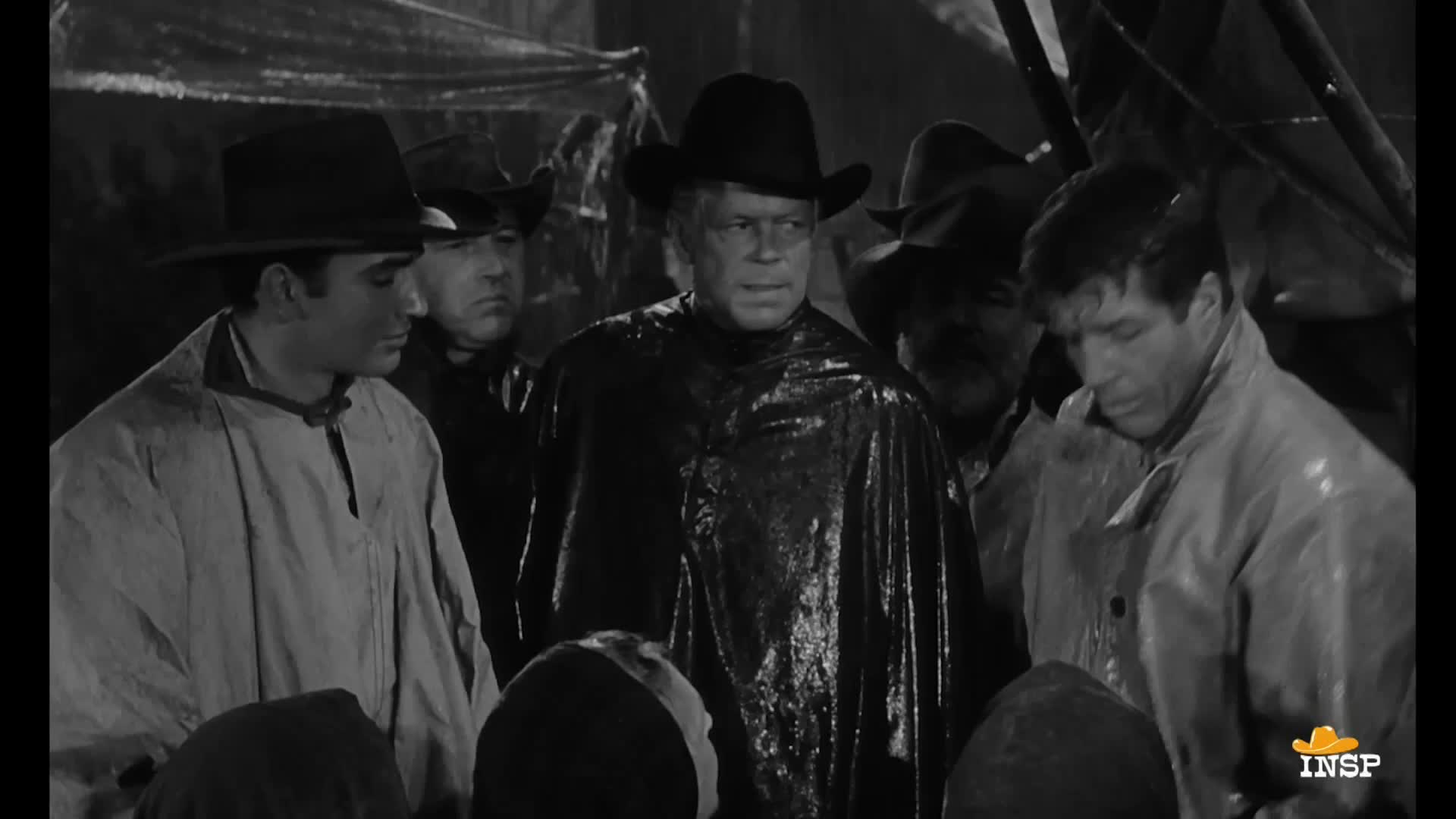 A group of men, all wearing hats and raincoats, stand close together in a dimly lit space. The man in the center, draped in a shiny black slicker, stares directly ahead.
