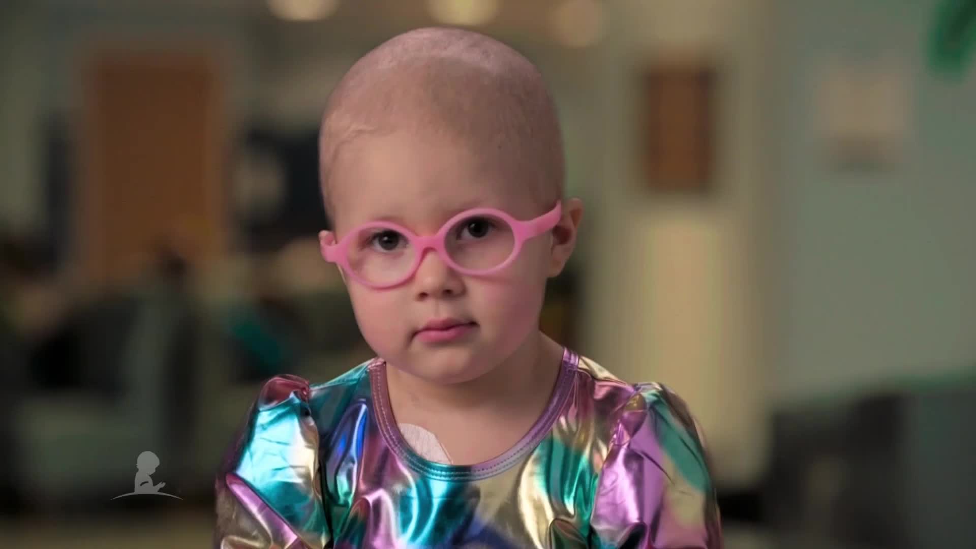A young child with a shaved head, wearing pink glasses and a colorful top, looks directly at the camera. The setting appears to be a hospital or clinic, possibly in the United States, as suggested by the INSP logo.

