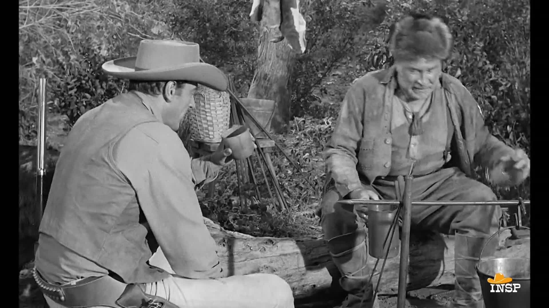 A cowboy, hat on, sits with a mug, while the older man stirs something in a pot over a fire. The scene, from an INSP show, is set outdoors, with trees and brush in the background.
