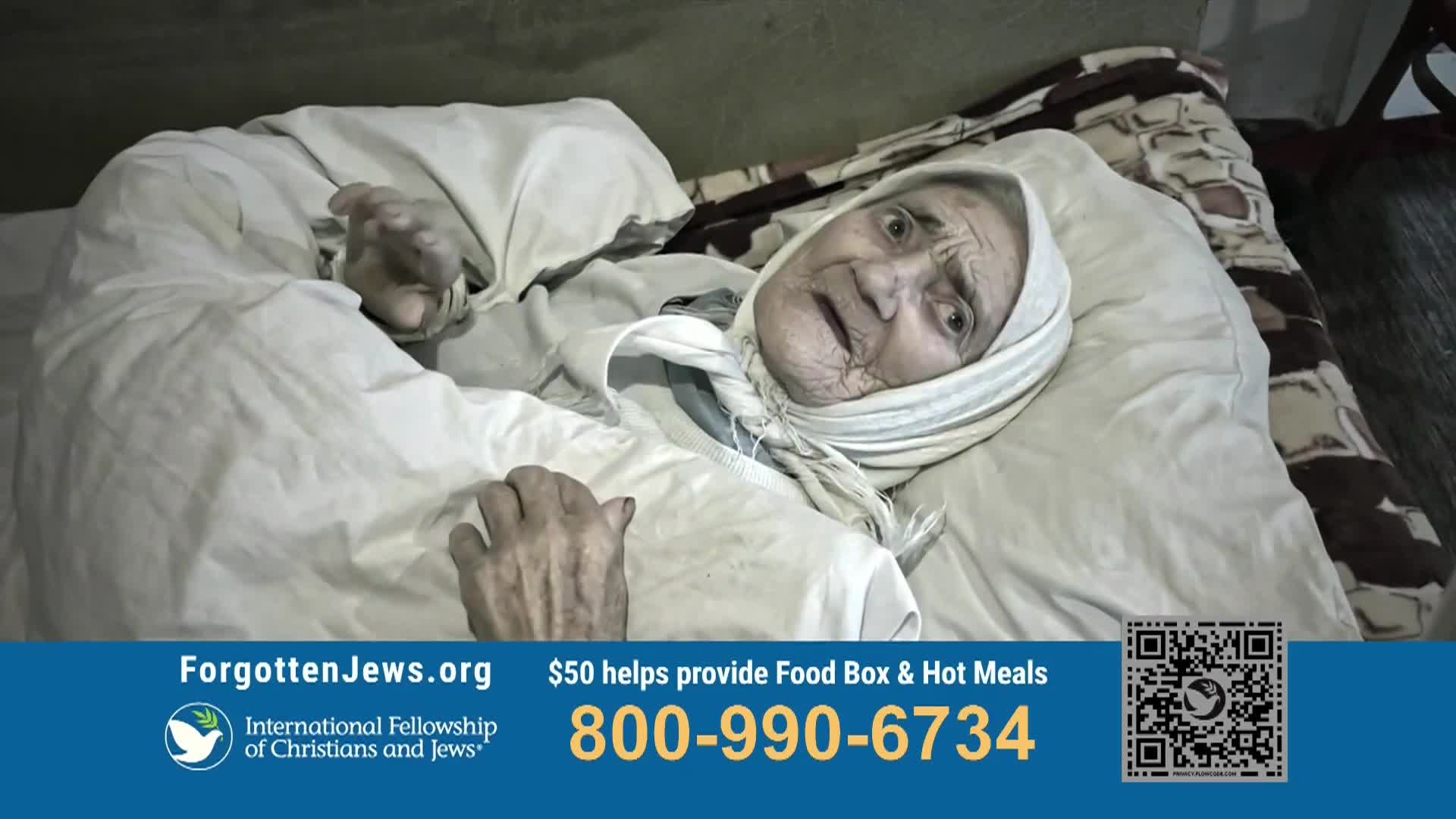 An elderly woman lies in bed, her eyes wide and hands slightly raised. She is wrapped in a blanket and wears a headscarf, with a look of concern on her face.
