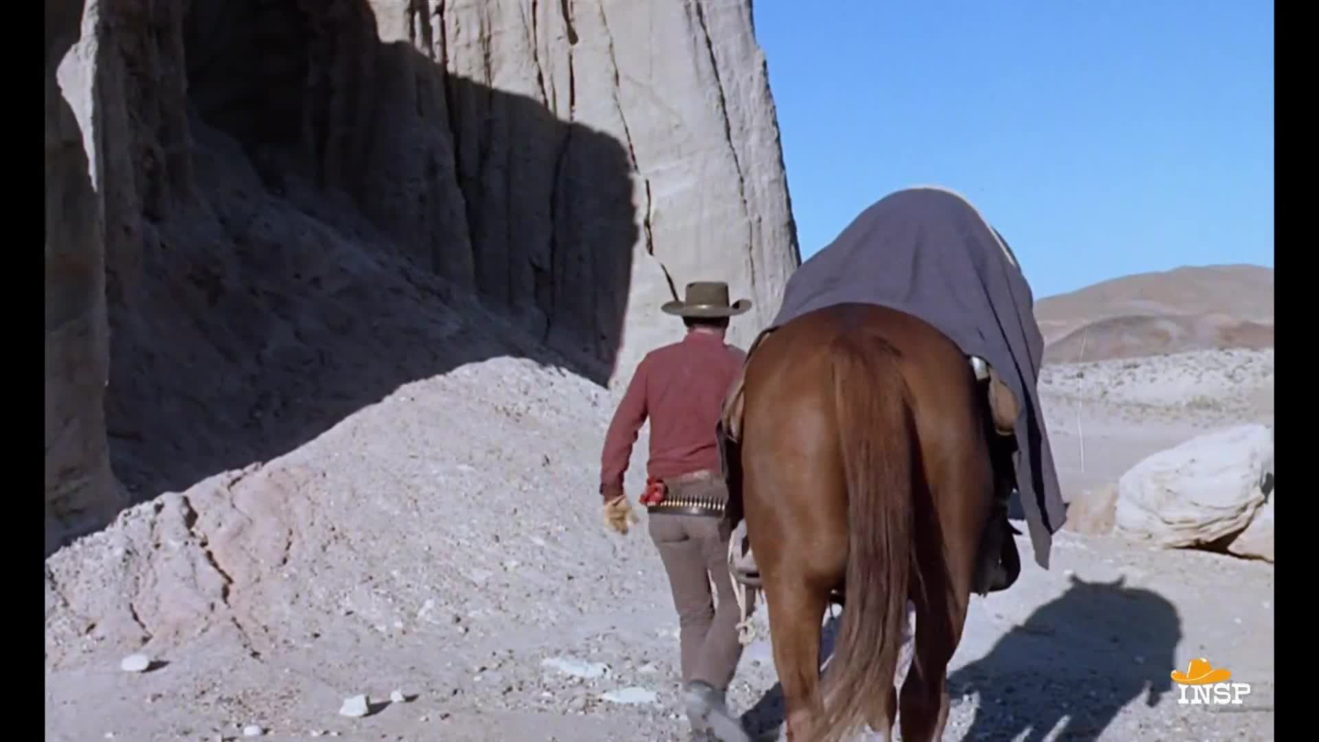 A cowboy in a red shirt and hat walks alongside his horse, leading it through a desert landscape. The horse is draped with a dark blanket, and the man's shadow stretches across the dusty ground.
