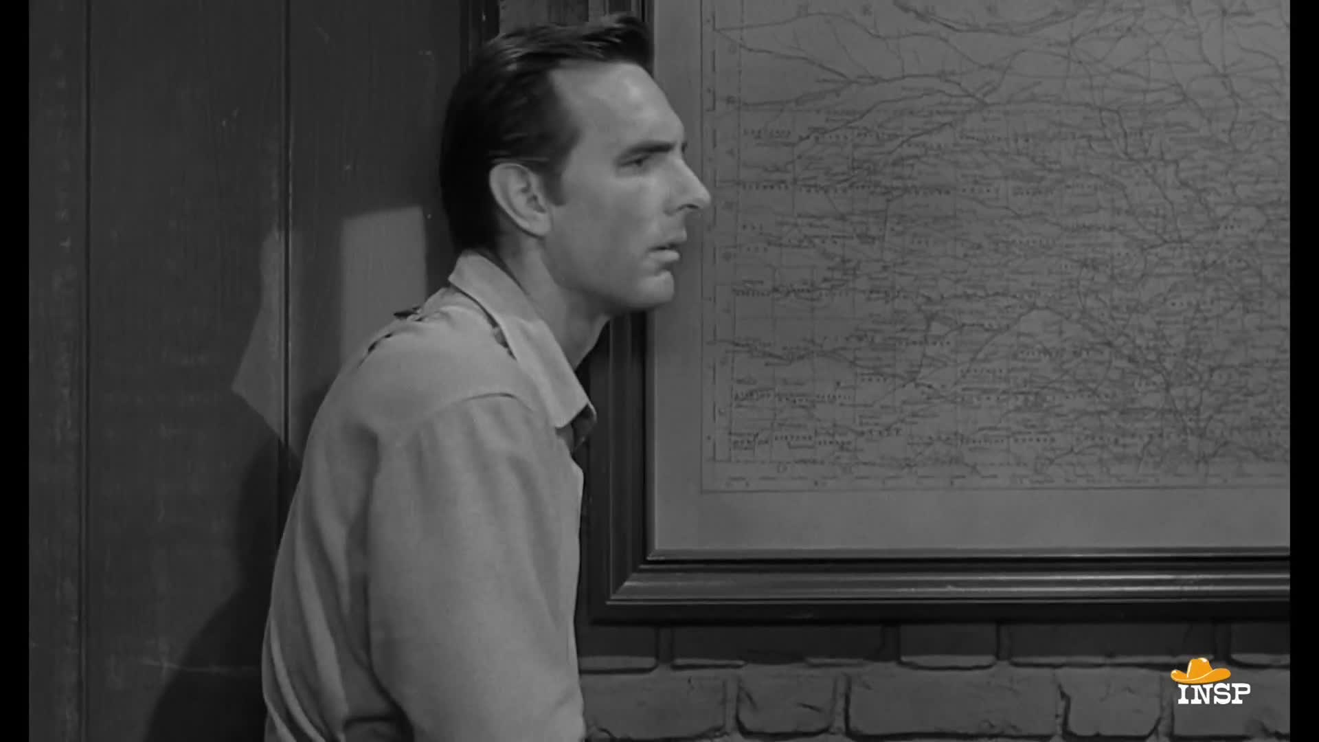 A man in a light-colored shirt leans against a wooden wall, his gaze fixed on a framed map. He seems lost in thought, his profile illuminated by the soft light.
