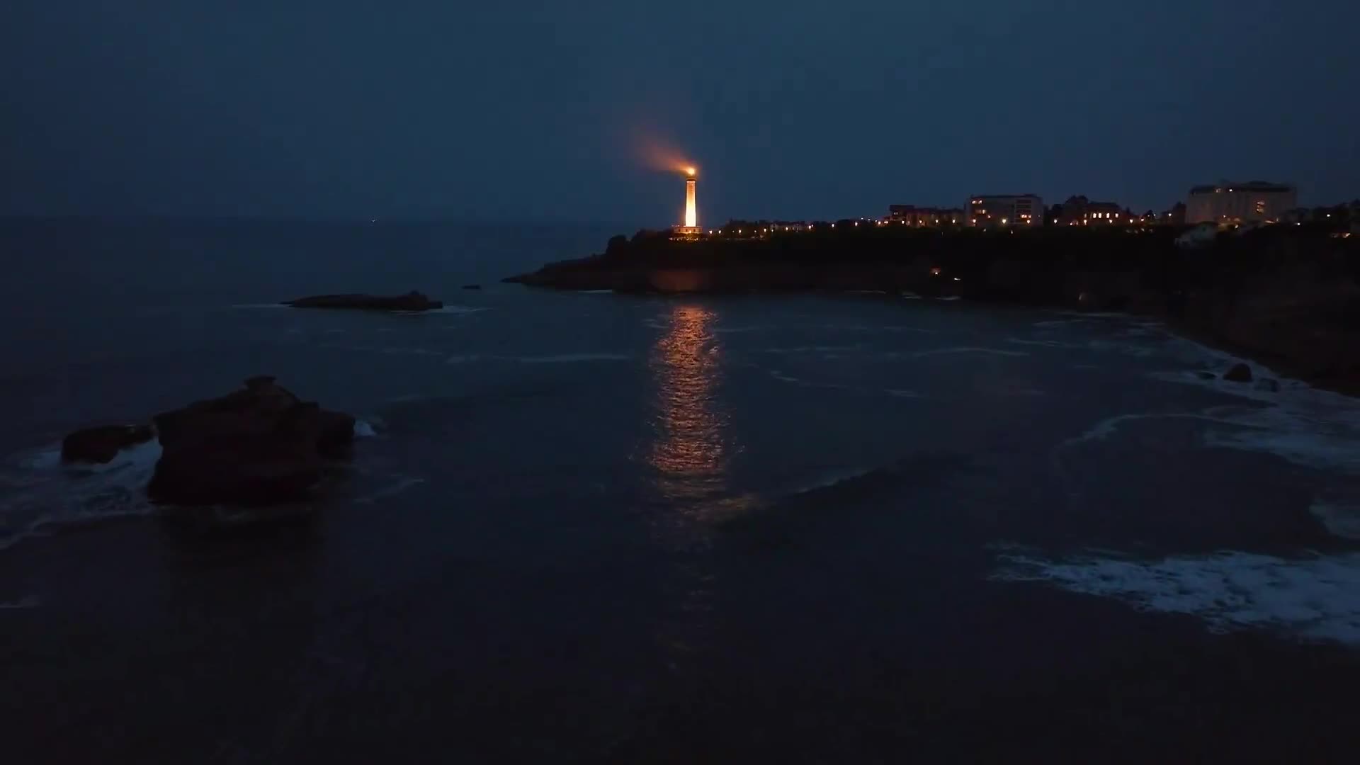 A lighthouse beam cuts through the dark sky, its reflection shimmering on the water. The waves gently lap against the shore, illuminated by the distant glow.
