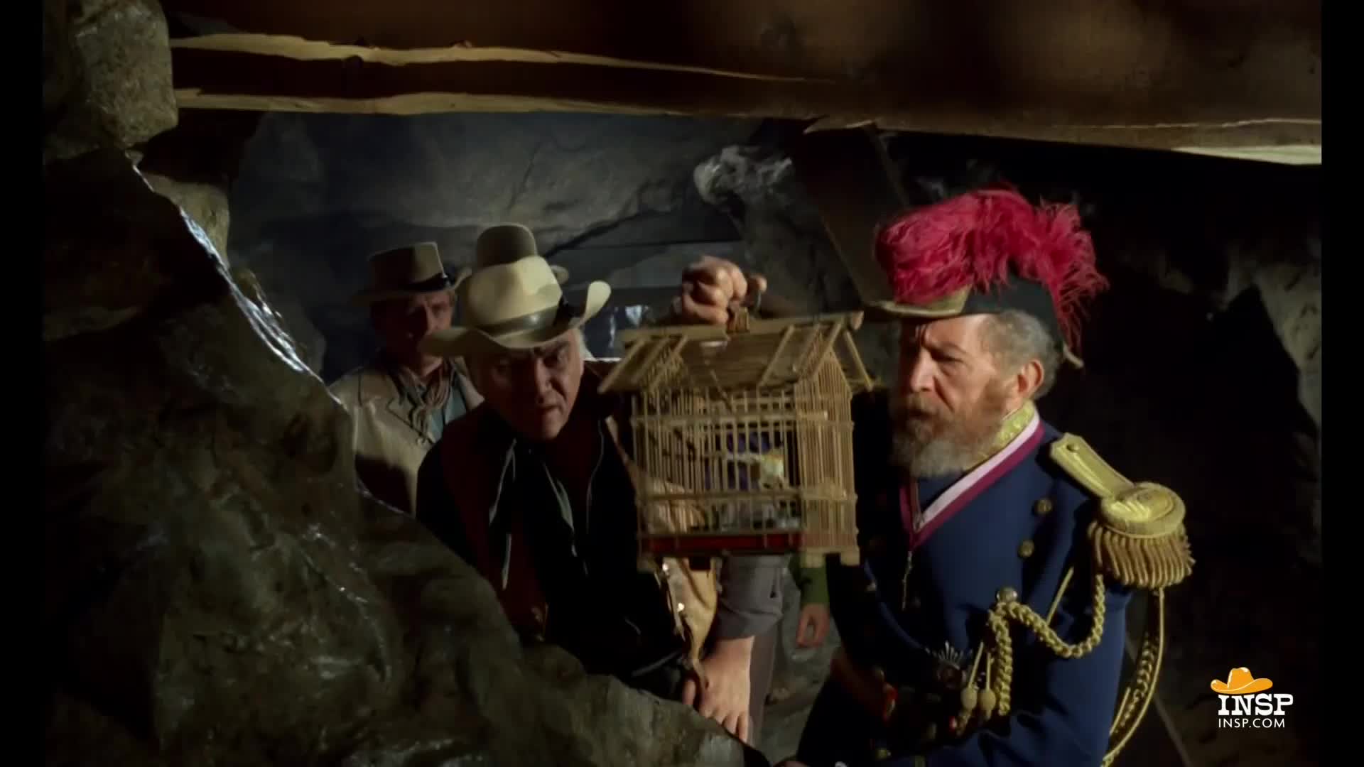 A man in a cowboy hat holds a birdcage, while a distinguished gentleman in a blue uniform and a red plume looks on. The group appears to be inside a cave or cavern.
