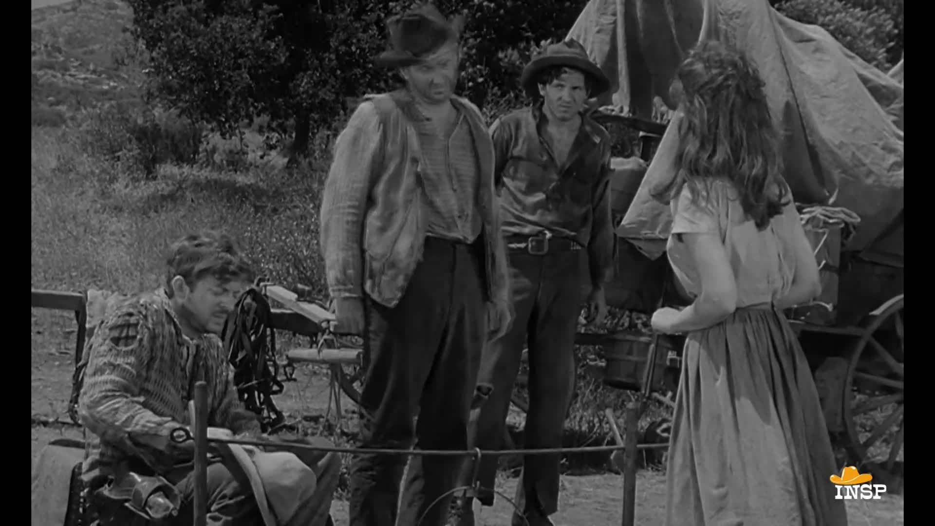 A woman faces the wagon, talking to the men standing nearby. One man sits, working on something with his hands, while the others look on.
