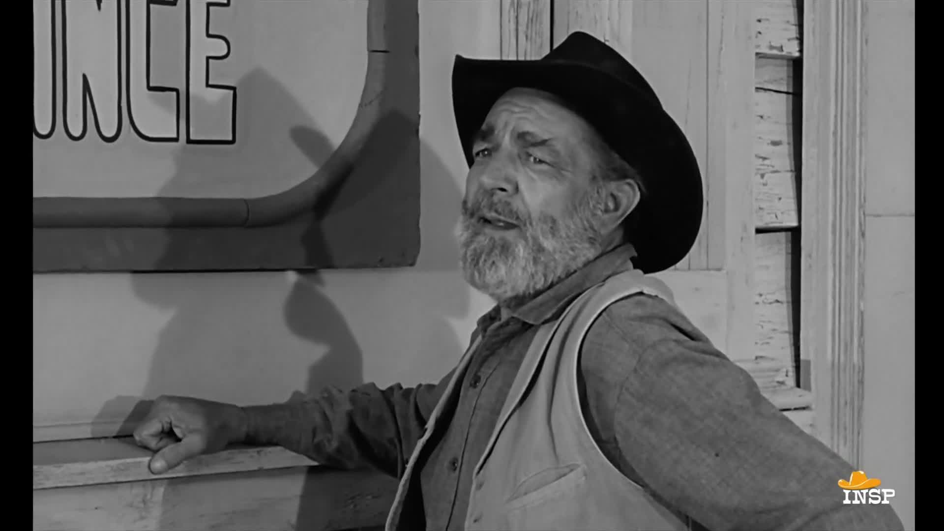 The man, wearing a hat and vest, leans against a weathered surface, his gaze directed upward. His hand rests casually on the ledge, and he seems to be in mid-conversation.
