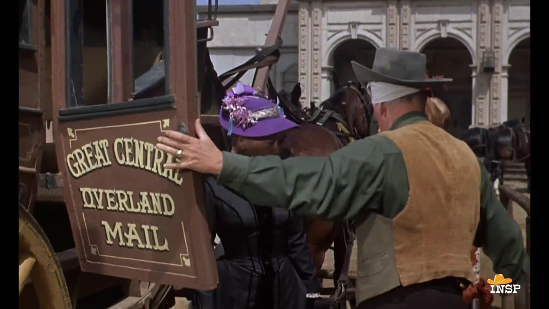 A man in a vest guides a woman, who's wearing a purple hat, towards a stagecoach marked "Great Central Overland Mail." The horses are hitched and ready to go.

