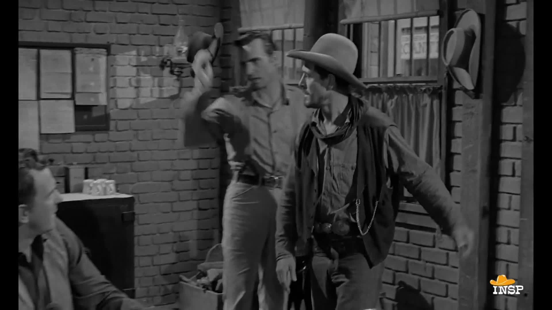 A man in a cowboy hat strides forward, his hand outstretched as if he's about to say something. Another man, standing behind him, holds a hat, looking on.
