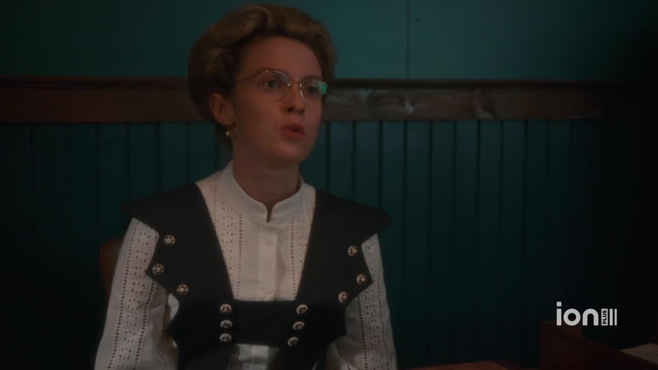 A woman in spectacles and a high-collared blouse speaks, her lips moving as she addresses someone off-screen. The ION Plus logo is visible in the bottom right corner.
