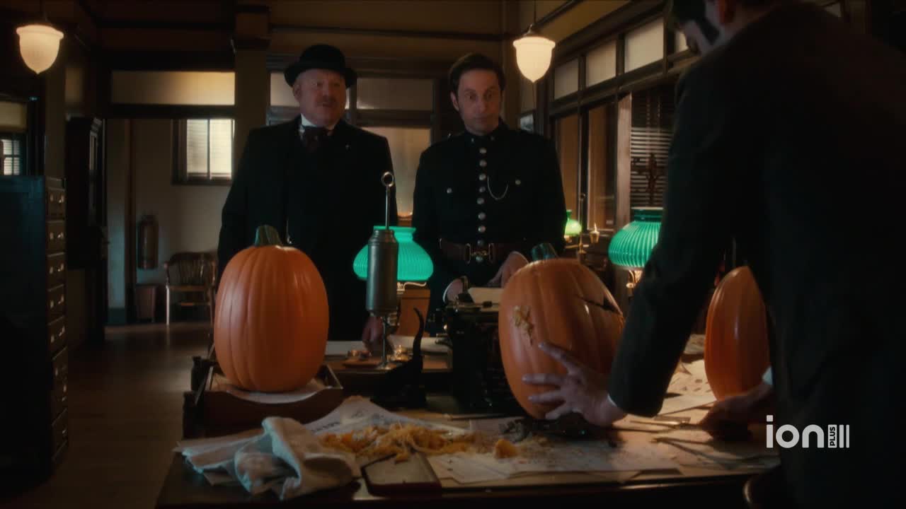 A man in a dark suit carves a pumpkin on a cluttered desk, while two other men in period attire watch. Pumpkin guts and seeds litter the surface, and the ION Plus logo is visible in the corner.
