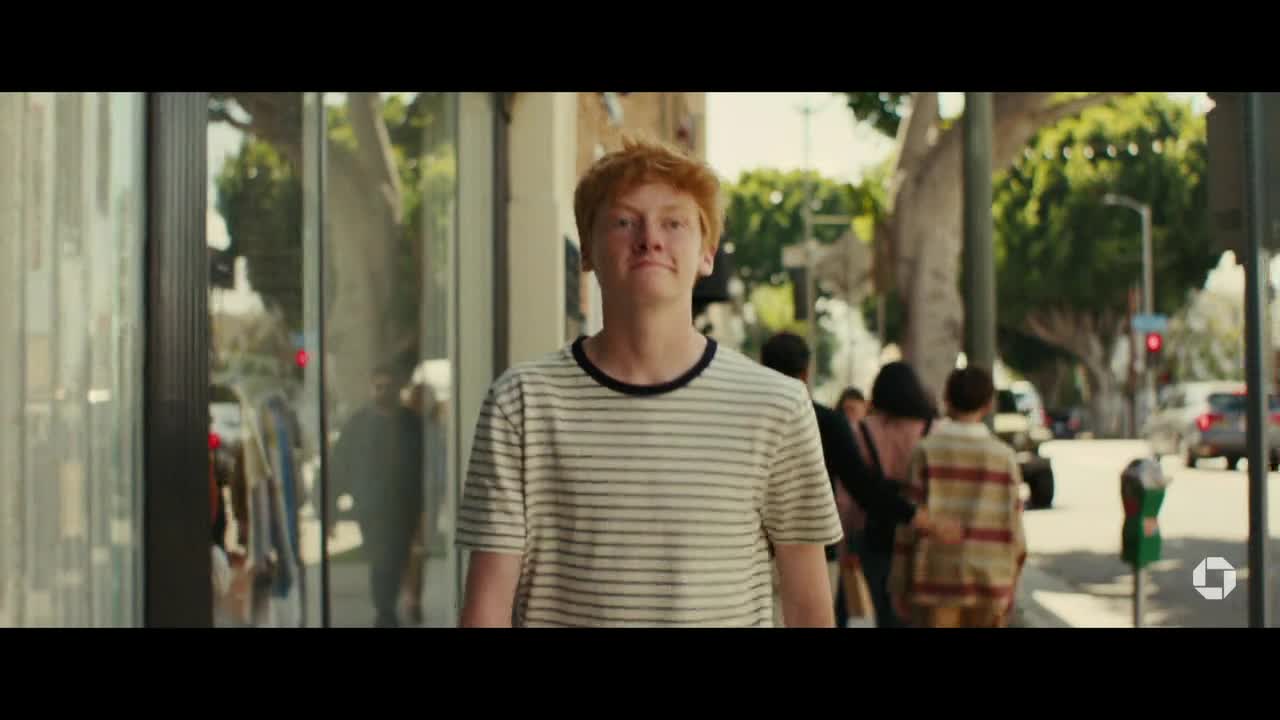 A young man with red hair in a striped shirt walks toward me on a sunlit sidewalk. Behind him, people stroll past storefronts and traffic moves on the street.
