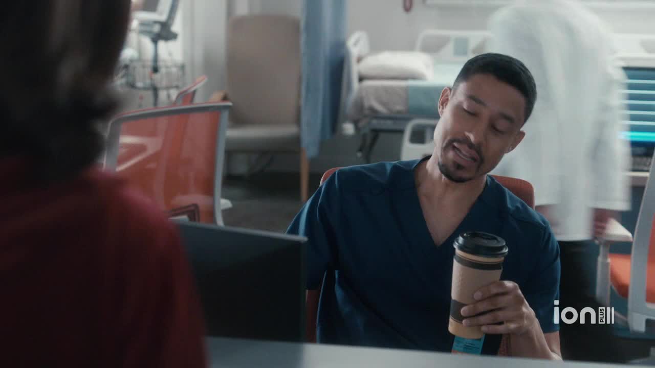 A man in blue scrubs leans back in his chair, holding a coffee cup. Someone in a white coat walks past in the background, near a hospital bed.

